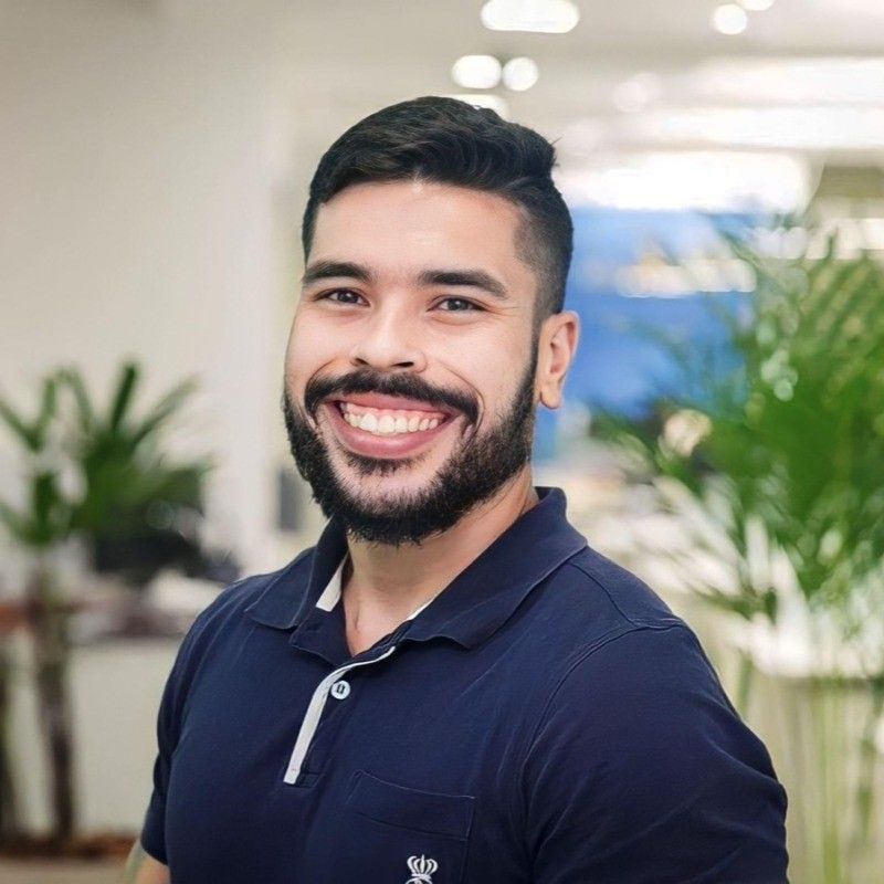 Homem de cabelos e barba escuros, sorrindo. Vestindo uma camisa polo azul-marinho em ambiente interno cercado por vegetação.