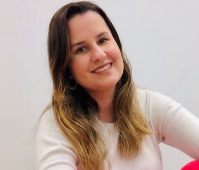 Mulher sorridente, vestindo um suéter branco, com longos cabelos castanhos. Sentada em frente a um fundo branco.