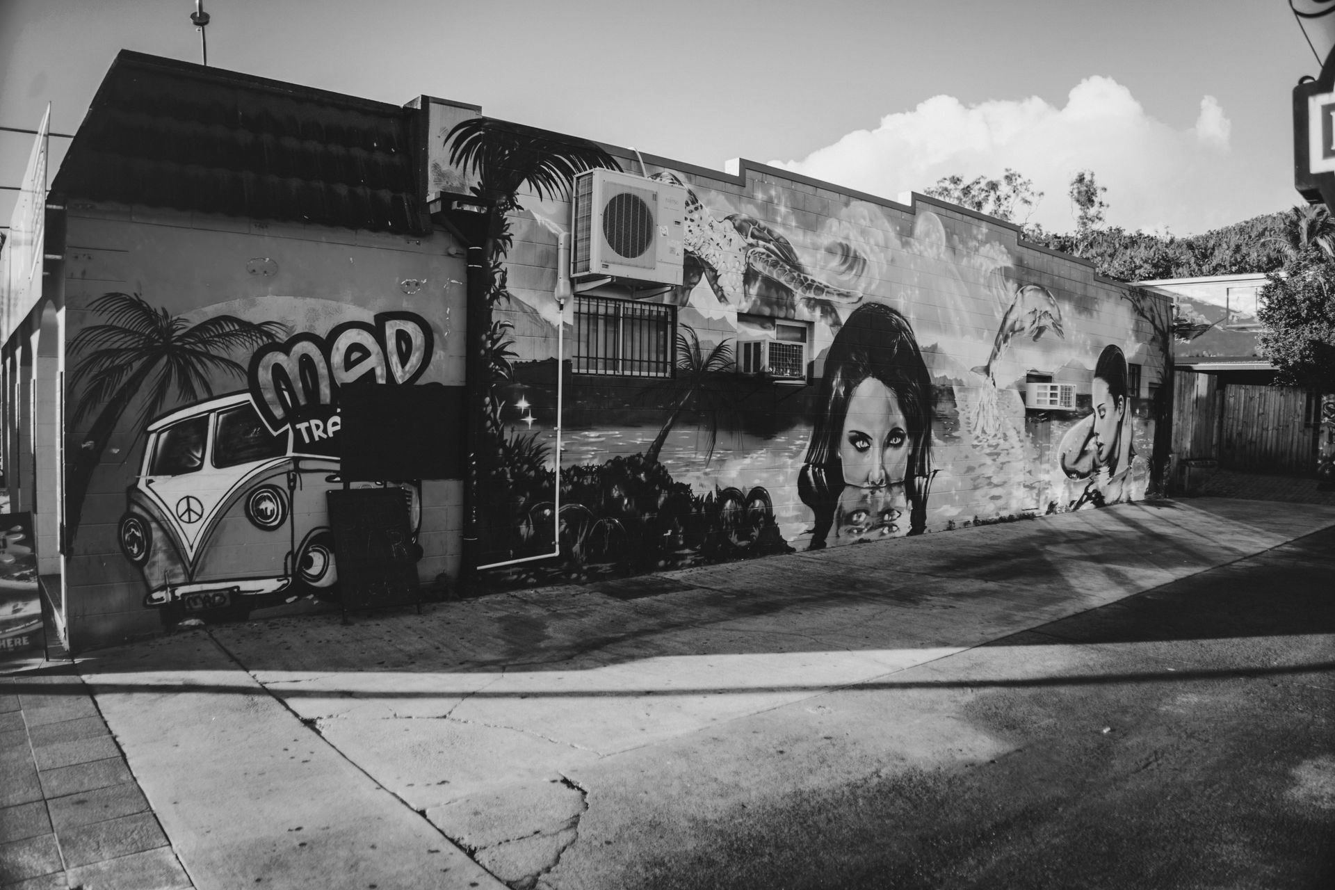 Mural em preto e branco na parede de um prédio com uma van, um rosto e uma cena tropical em uma rua da cidade.