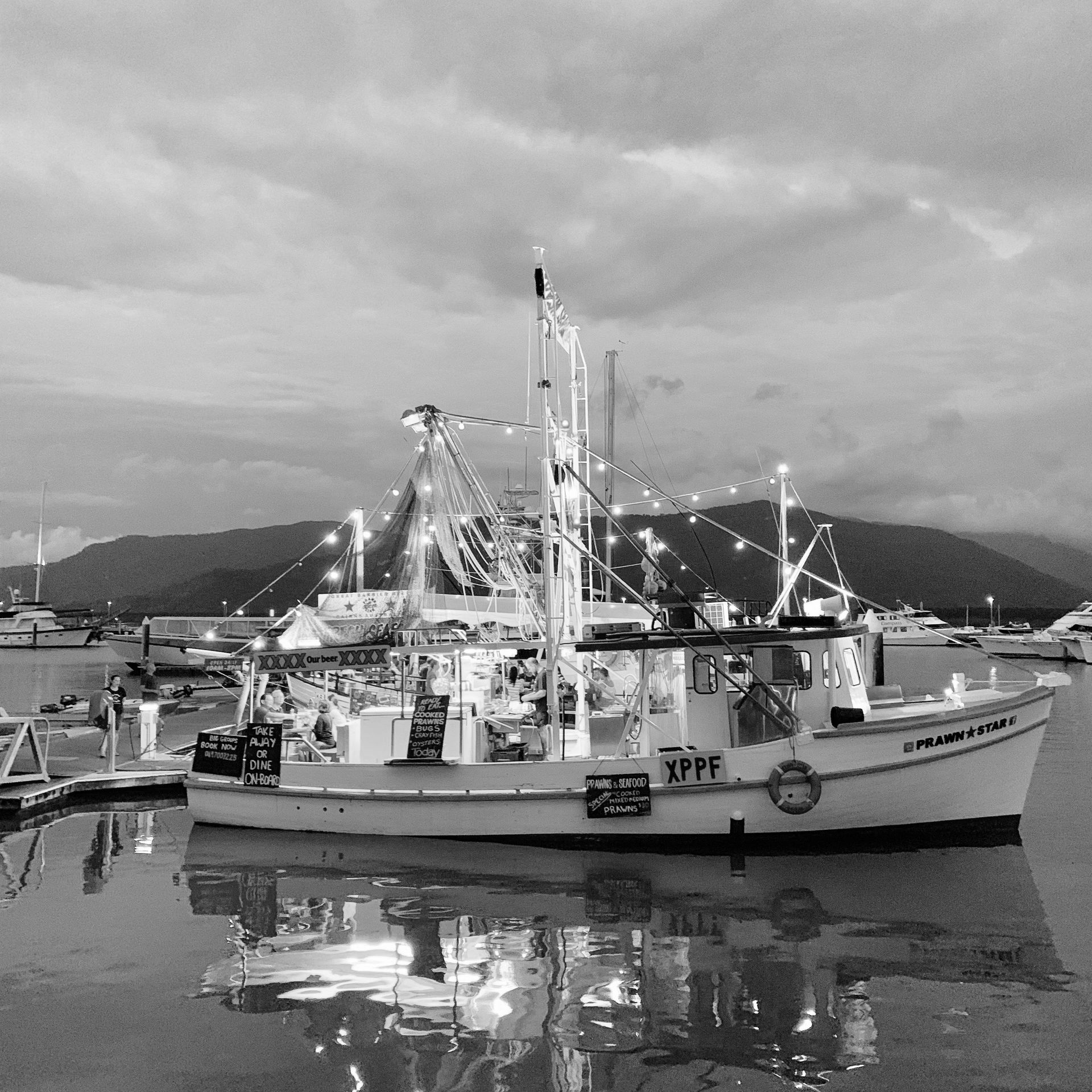 Barco de pesca iluminado com luzes em um porto, montanhas ao fundo, água refletindo as luzes e nuvens.