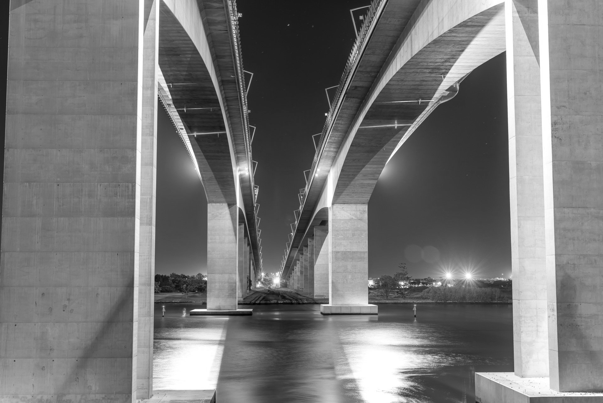 Foto em preto e branco de duas pontes elevadas à noite, atravessando um corpo d'água, com luzes.