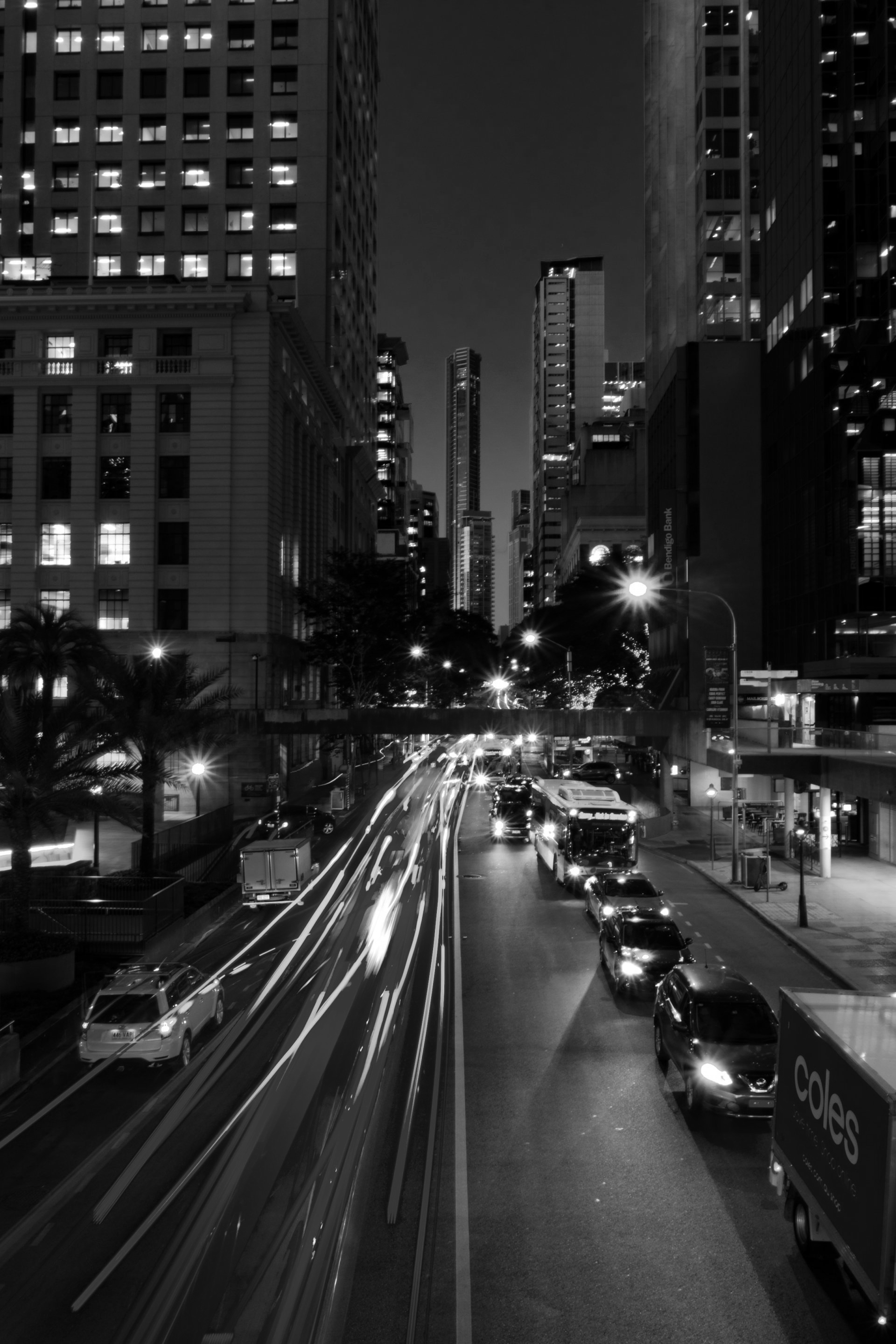 Rua da cidade em preto e branco à noite com faixas de luzes de carros e prédios altos.