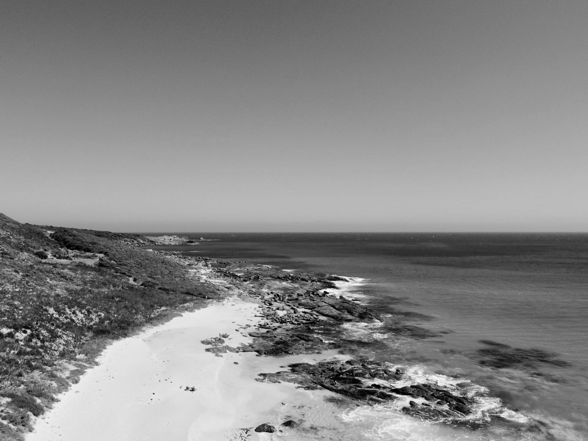 Foto em preto e branco de uma praia com pedras, vegetação e o oceano calmo sob um céu limpo.