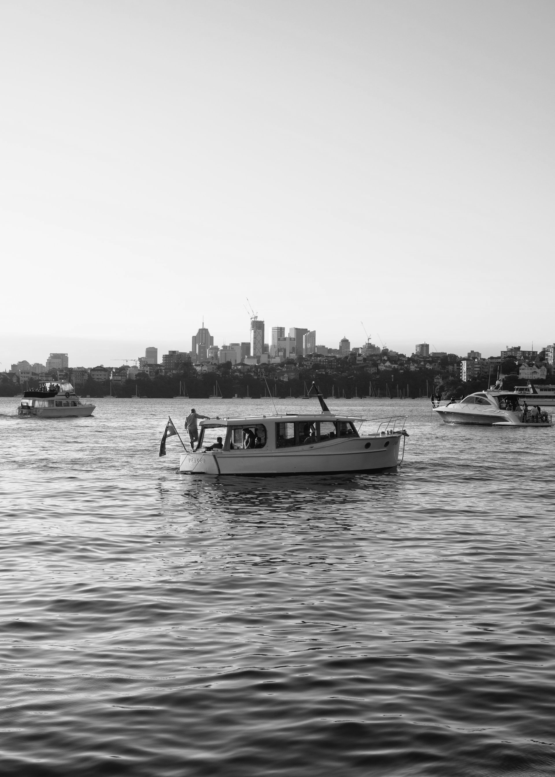 Barcos na água com o horizonte de uma cidade ao fundo; uma pessoa em pé no convés de um barco.