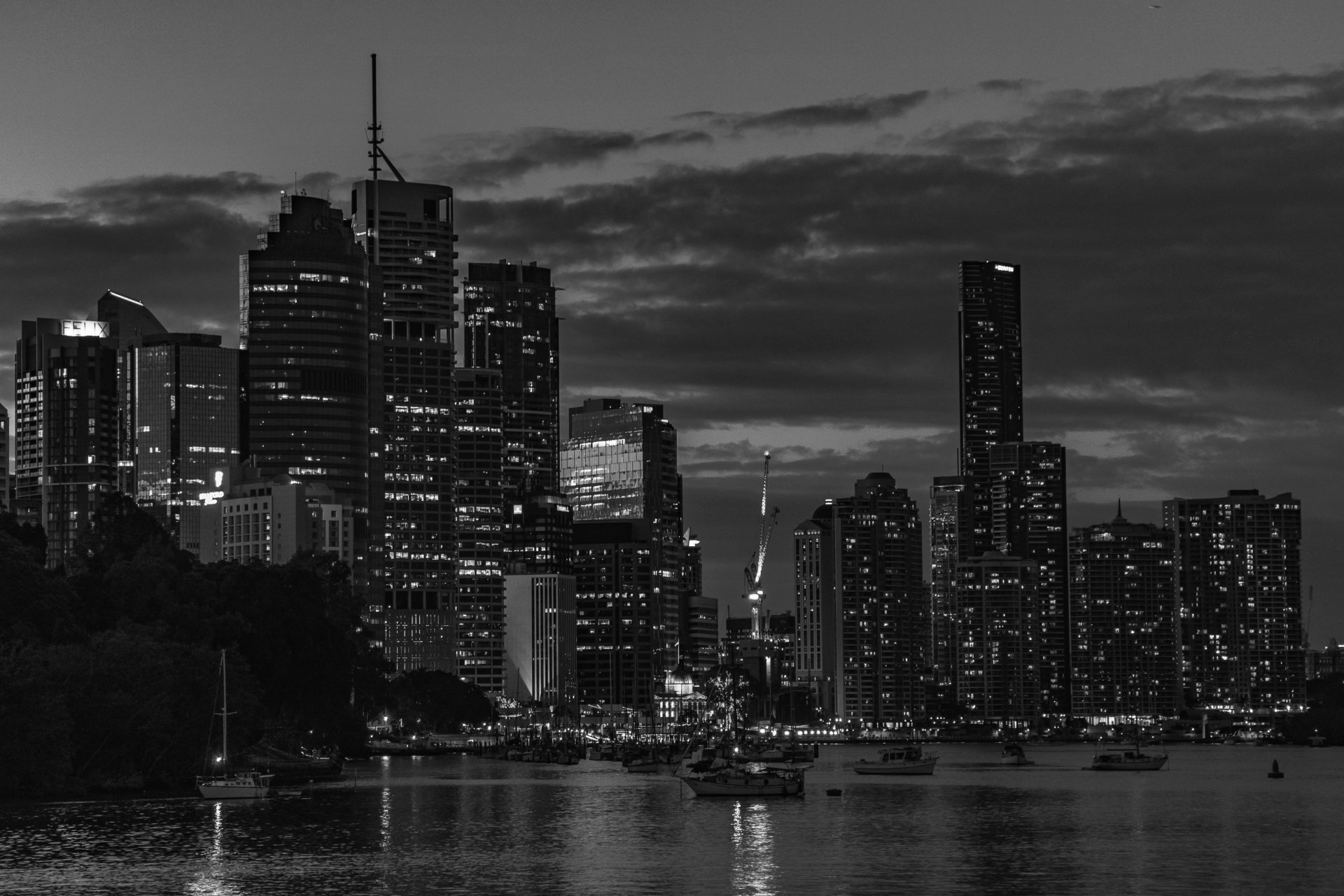 Paisagem urbana em preto e branco ao anoitecer, com edifícios iluminados refletindo na água e veleiros em primeiro plano.