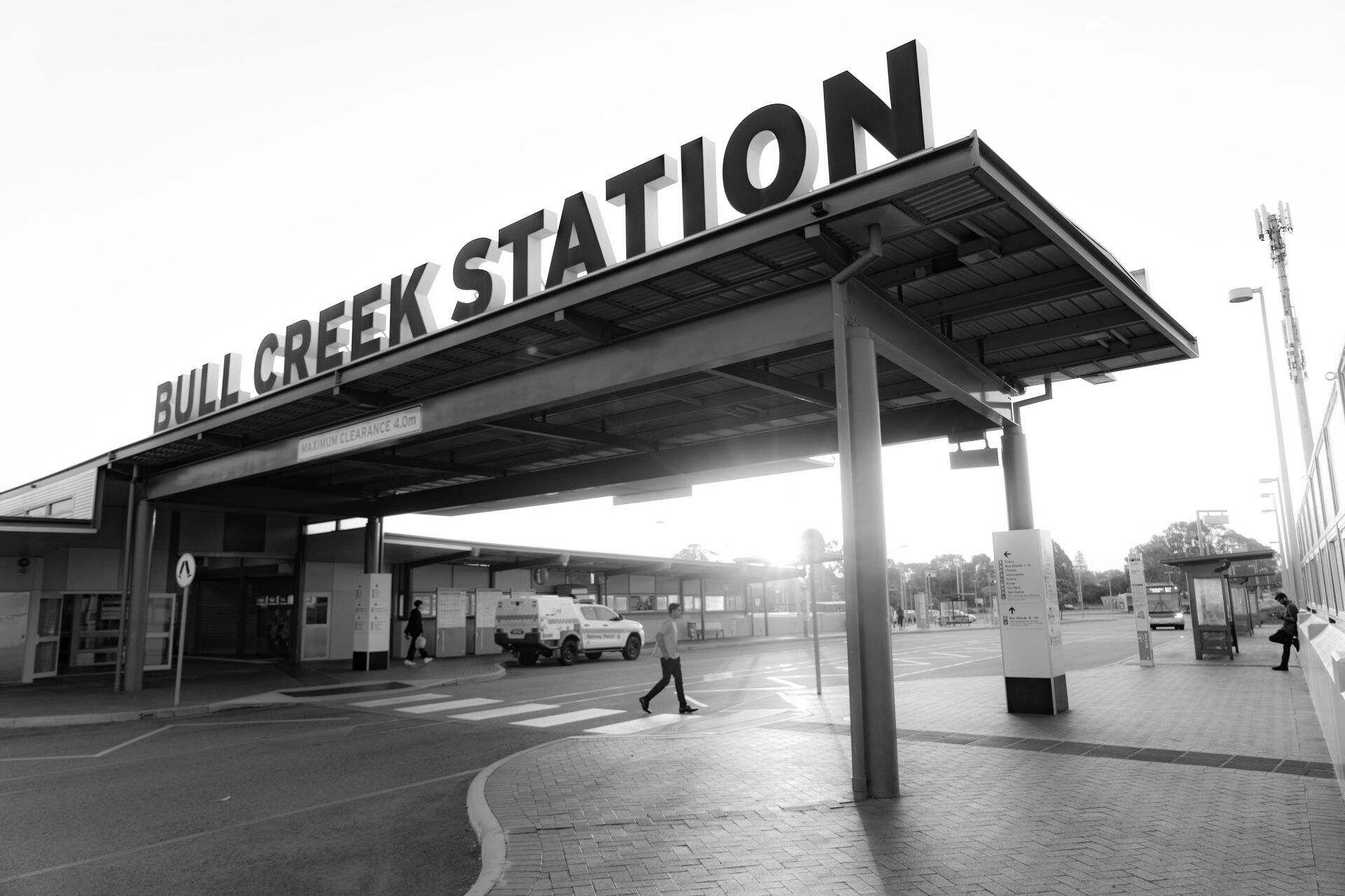 Entrada da Estação Bull Creek com placa, pessoa atravessando a rua, céu nublado, preto e branco.