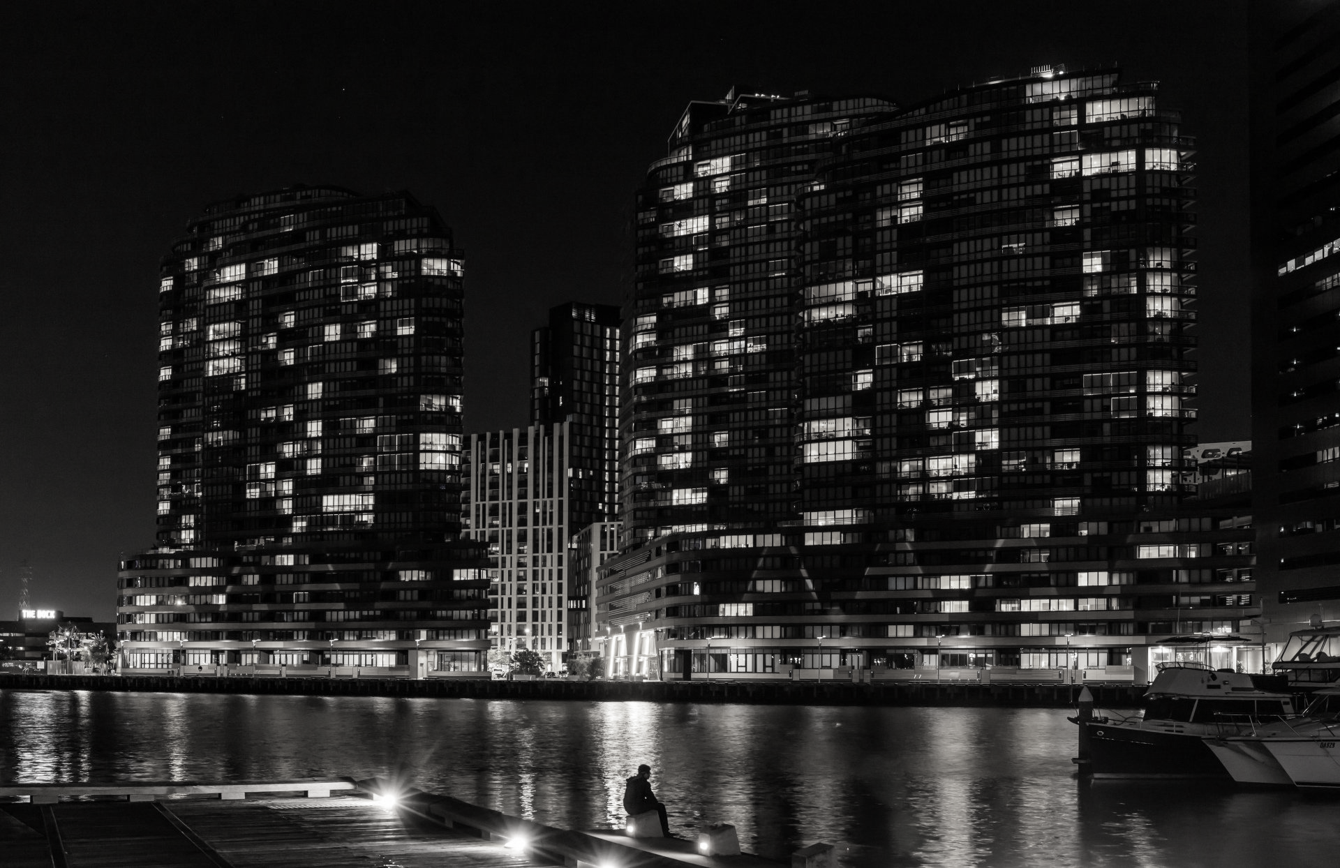 Fotografia em preto e branco de arranha-céus iluminados à noite refletidos na água, com uma pessoa sentada em um cais.