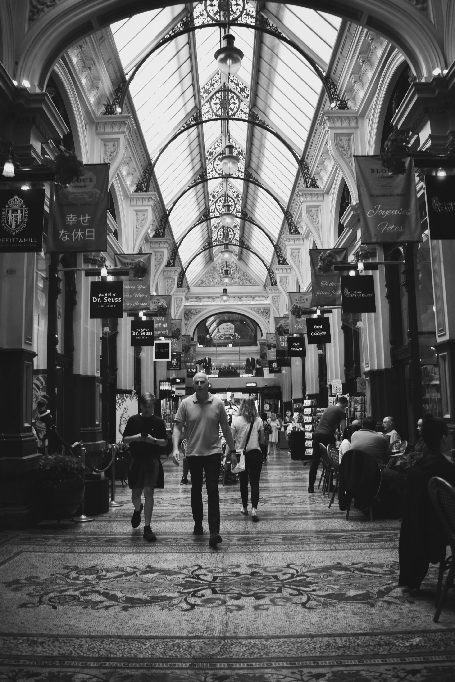 Pessoas caminhando em uma galeria comercial iluminada, com arquitetura ornamentada e teto de vidro.