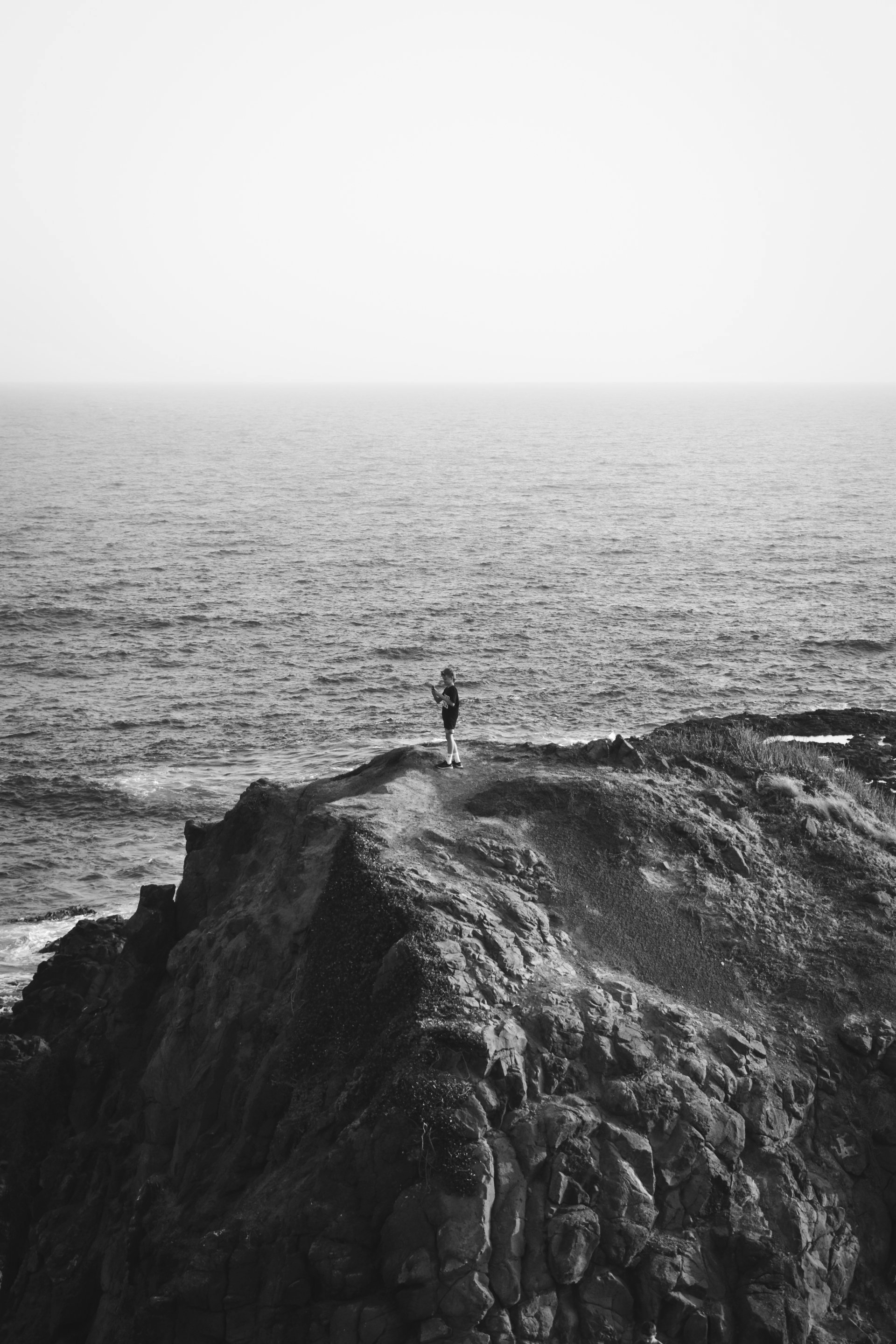 Pessoa em pé sobre um penhasco rochoso, com vista para o oceano. Céu nublado em preto e branco.