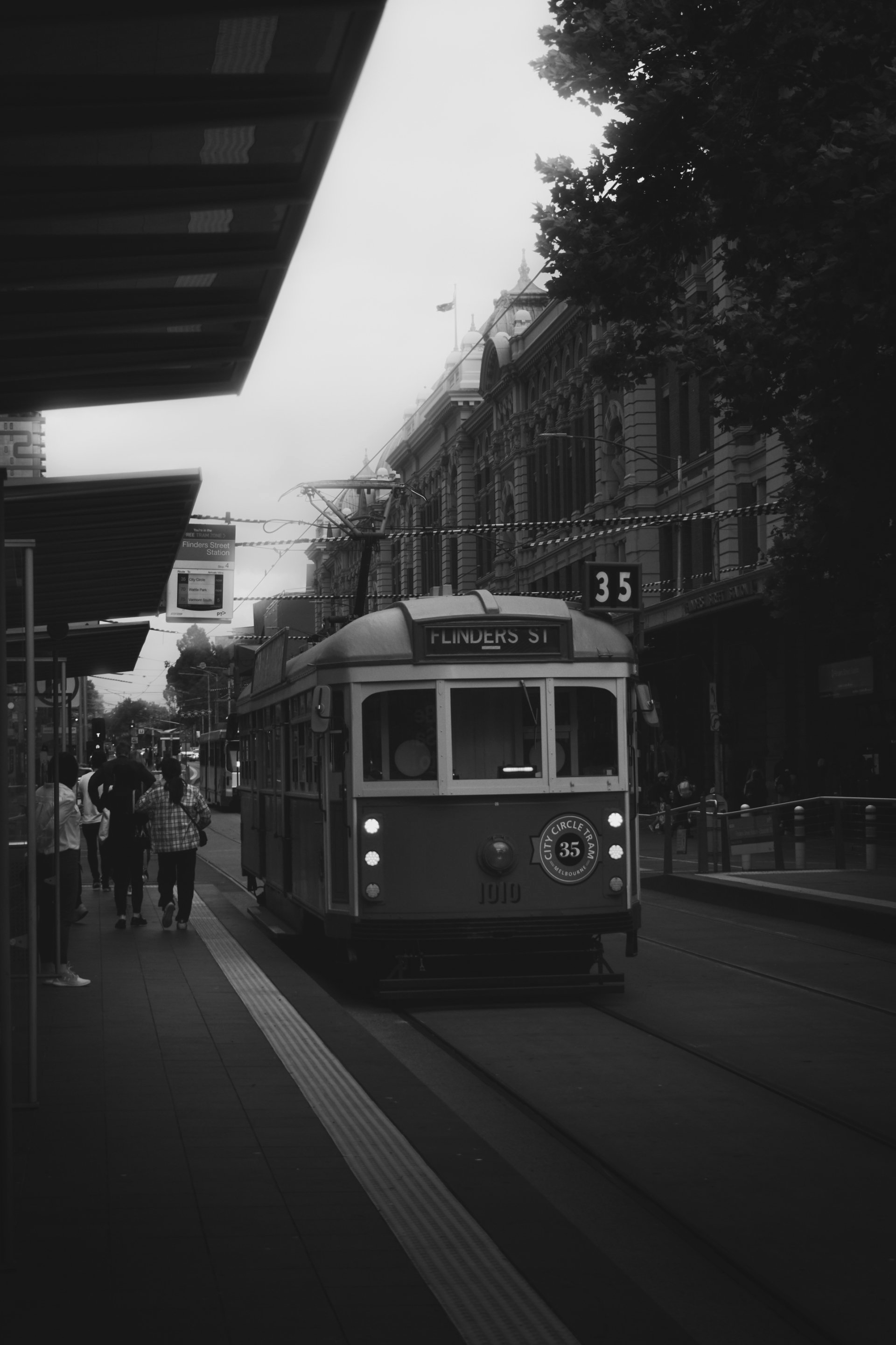Foto em preto e branco de um bonde sobre trilhos em um ambiente urbano com pessoas esperando em um ponto.