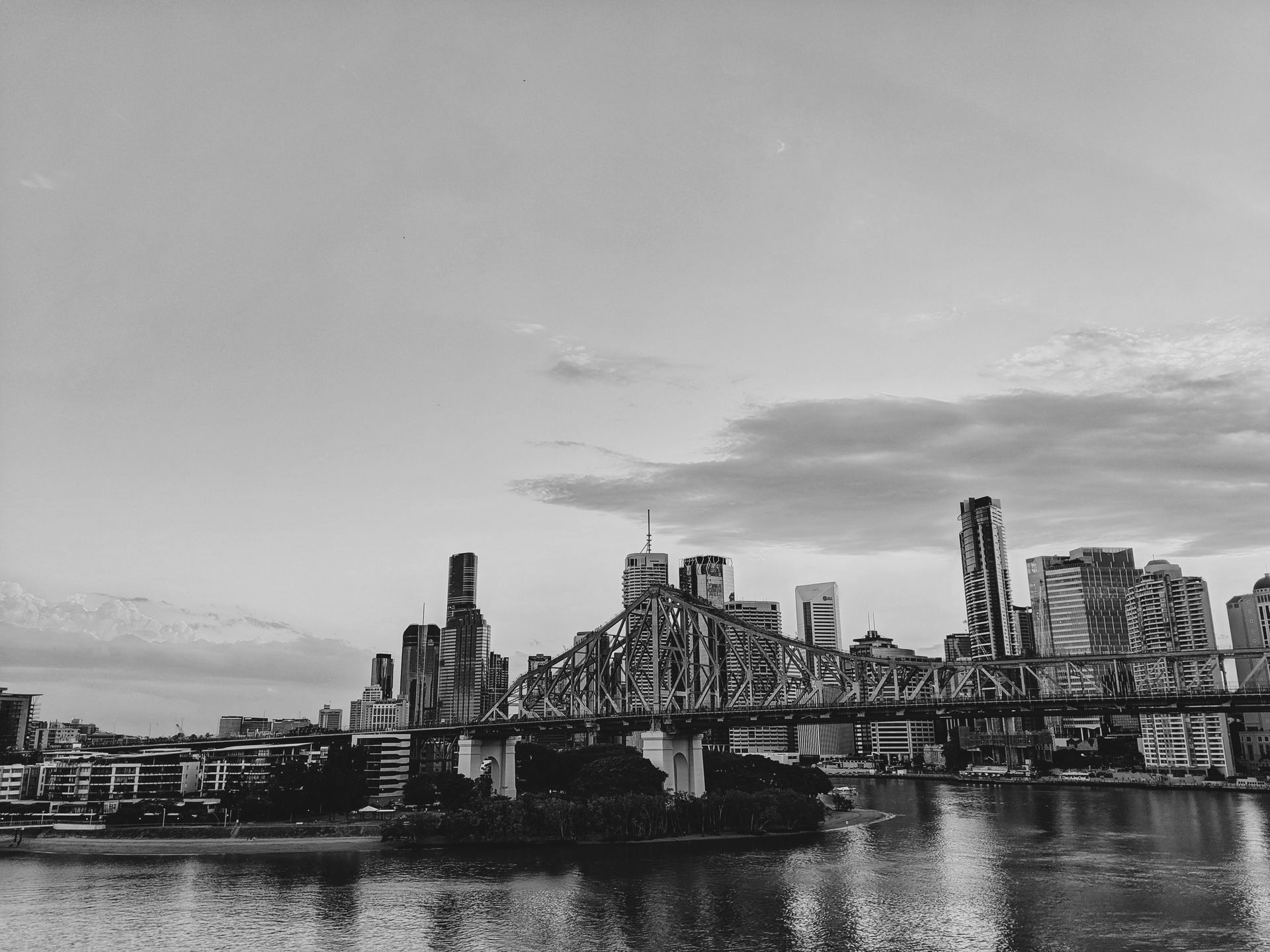 Vista em preto e branco do horizonte de uma cidade com uma ponte sobre um corpo d'água sob um céu nublado.