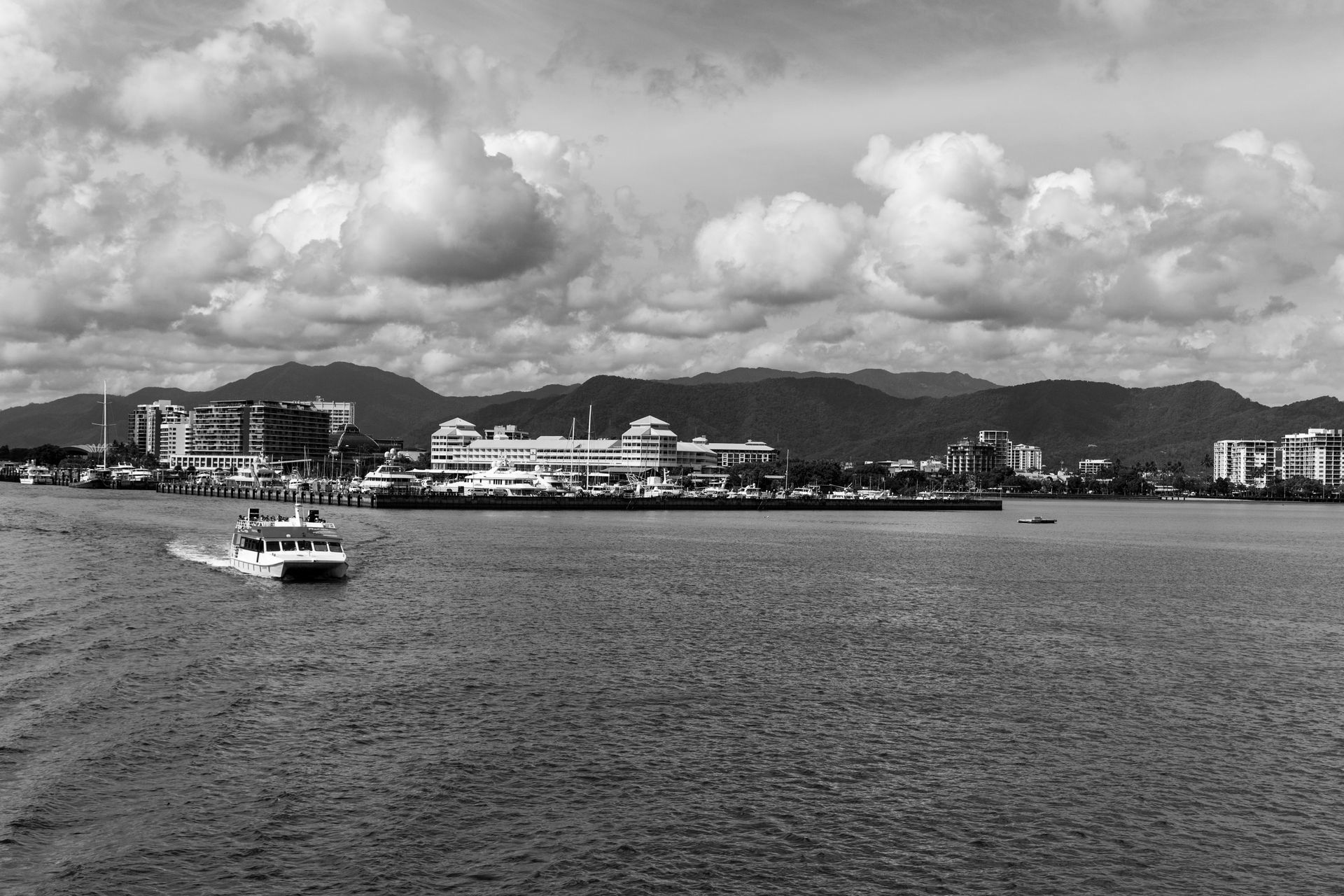 Vista em preto e branco de um porto com uma balsa, edifícios na costa e montanhas ao fundo.