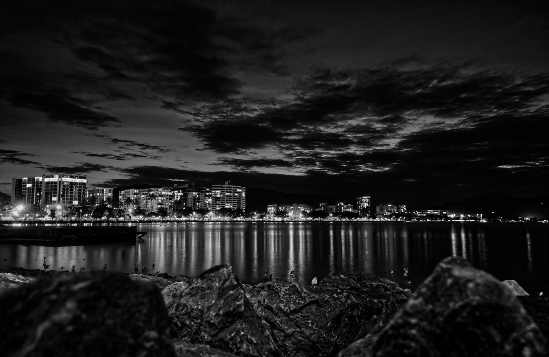 Horizonte da cidade à noite refletido na água, nuvens escuras no alto.
