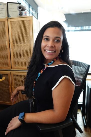 Uma mulher de longos cabelos escuros sorri, apoiando a bochecha na mão, sentada em uma cabine com encosto verde.