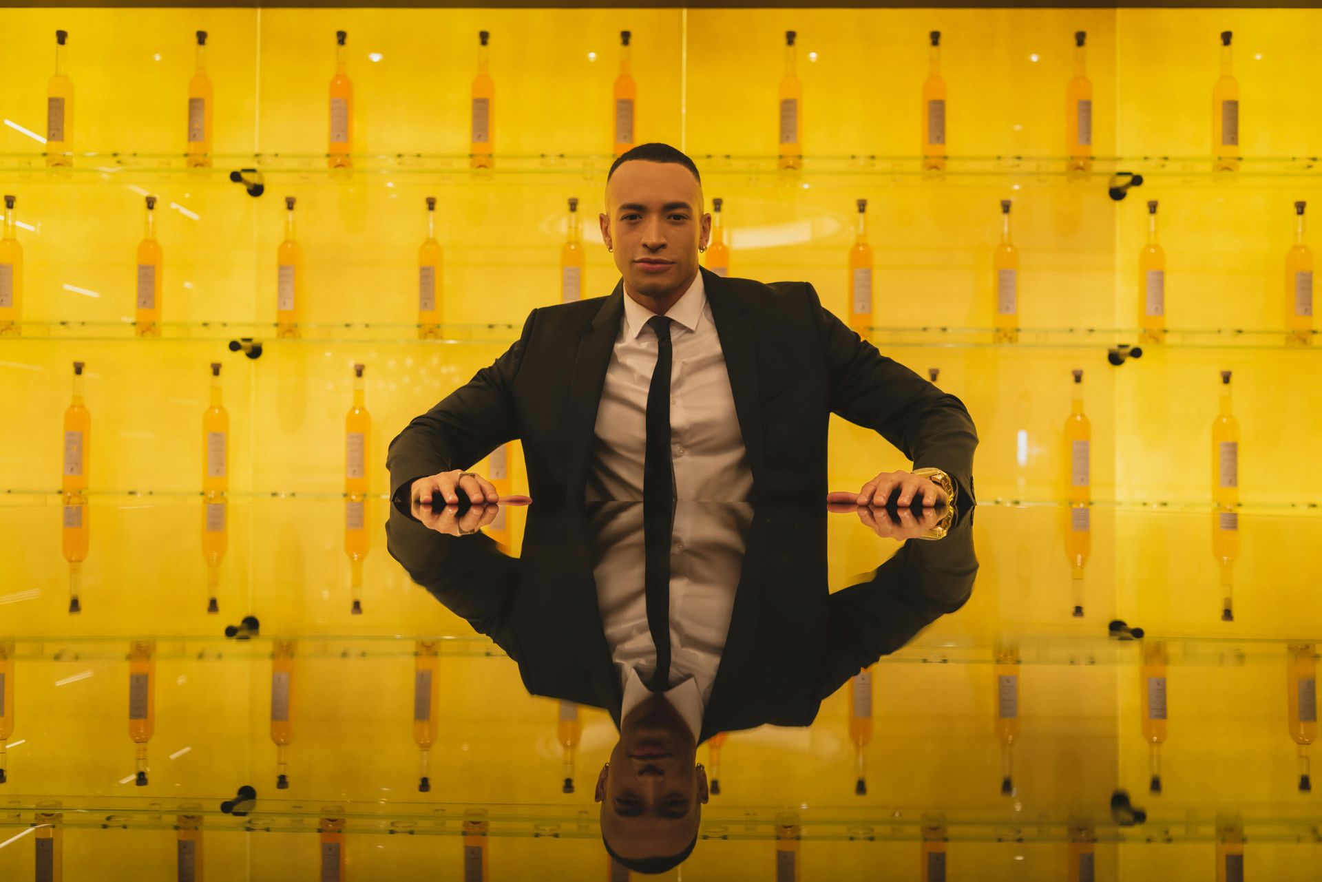 Man in a dark suit sitting against a bright yellow mirrored wall, centered in frame.
