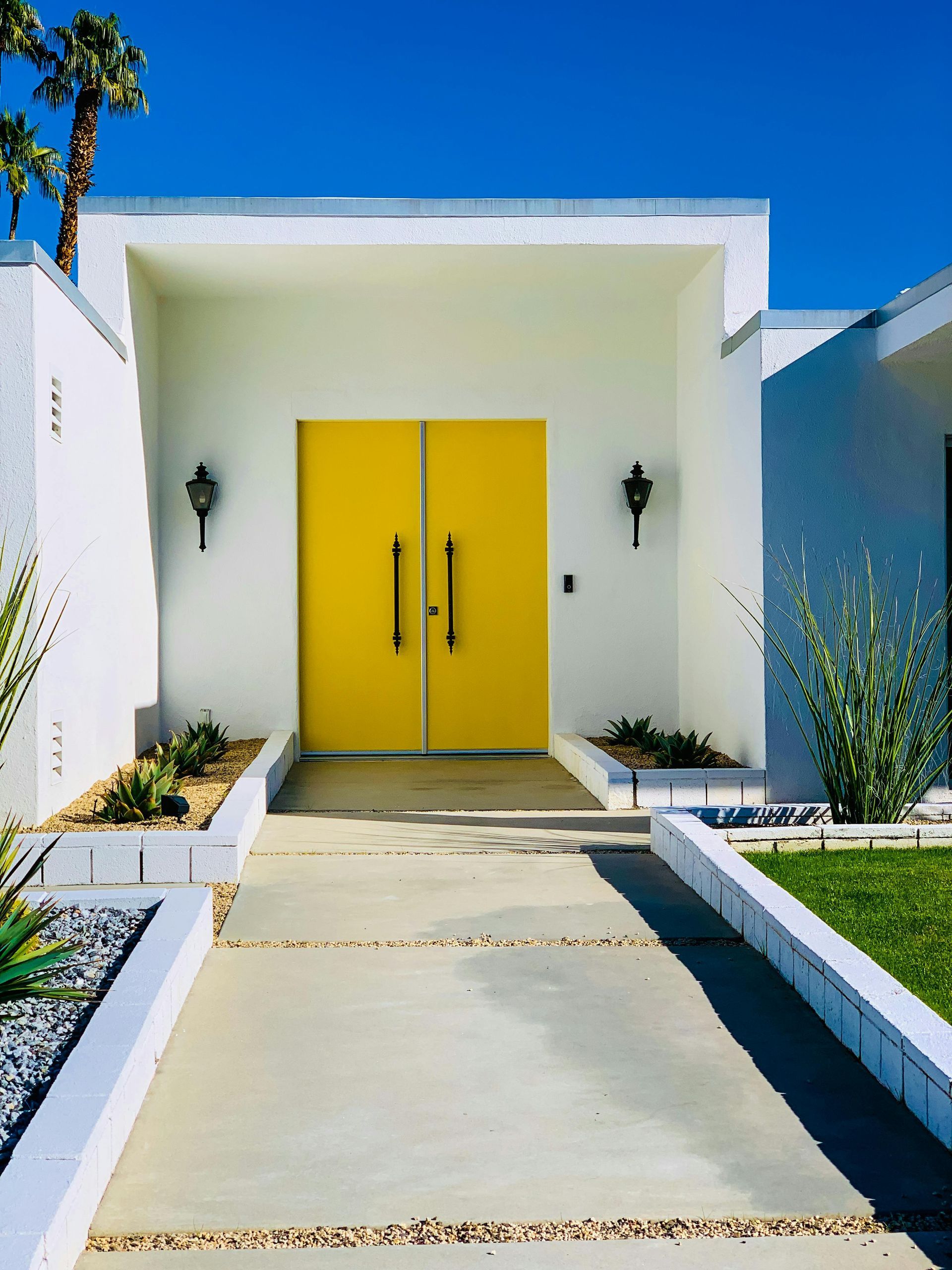 Modern white house entrance with bright yellow double doors and a concrete walkway, flanked by small garden beds