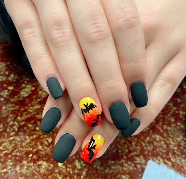 Hands with matte black nails, two accent nails feature a sunset design with bats.