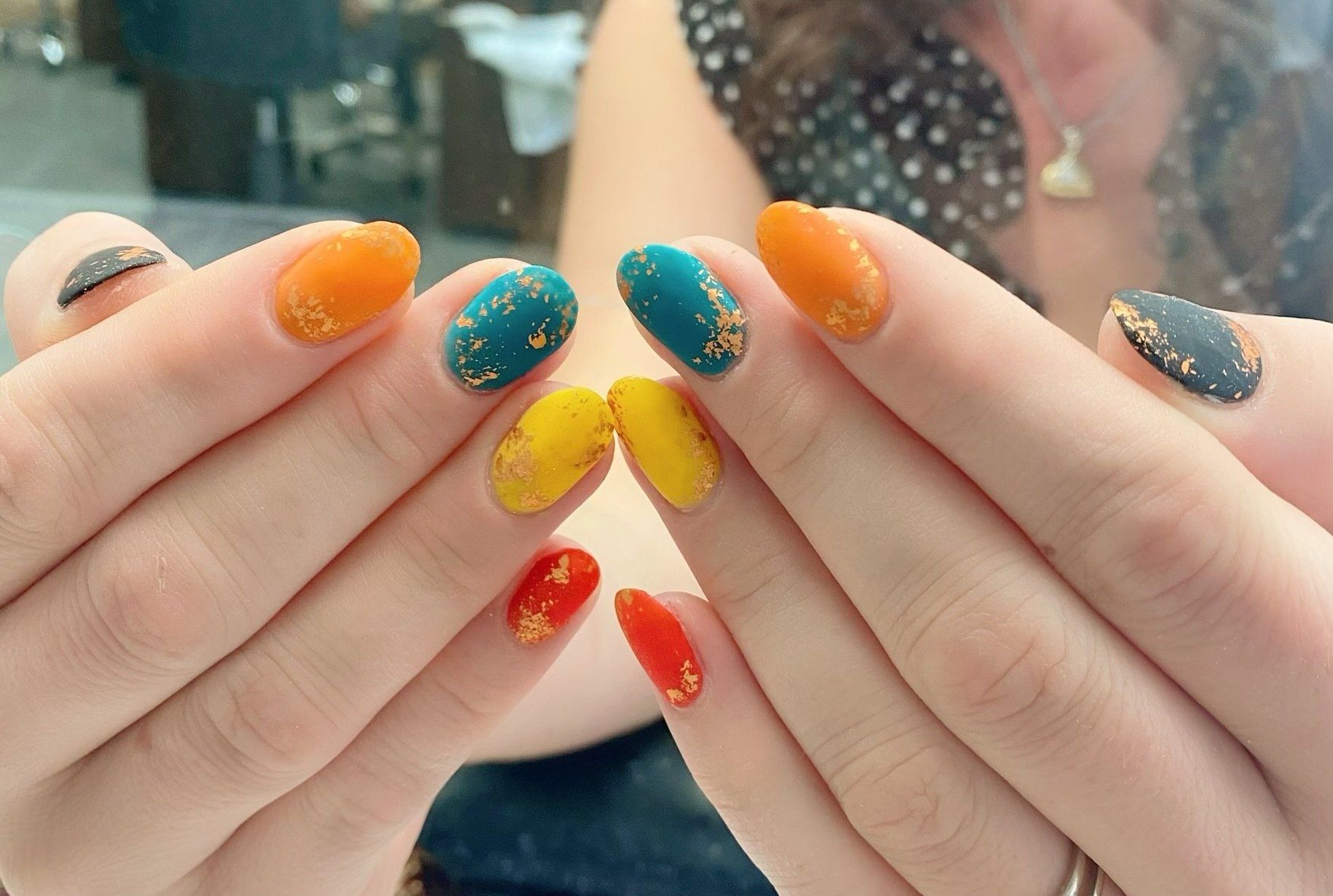 Hands displaying colorful nail art: orange, teal, yellow, red with gold flecks.