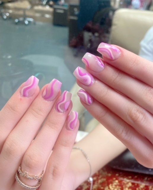 Hands with pink and white swirl design on square-shaped nails, a golden ring on a finger.