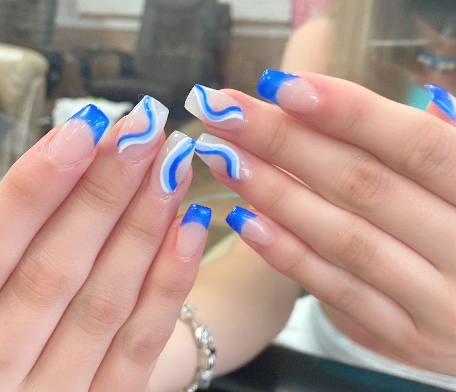 Hands with square, clear nails, blue tips, and white/blue wavy designs.