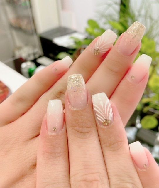 Hands with elegant square-shaped nails, ombre pink and gold glitter, some with feathery white designs and rhinestones.