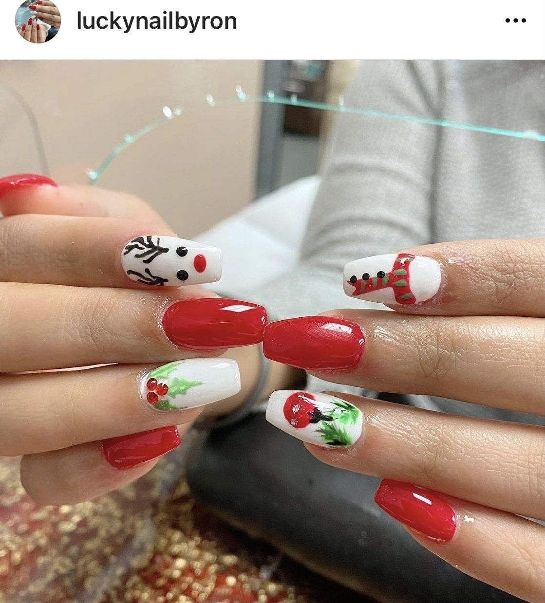 Christmas-themed nail art. Red and white polish with reindeer, boots, and holly designs. Hands in focus.