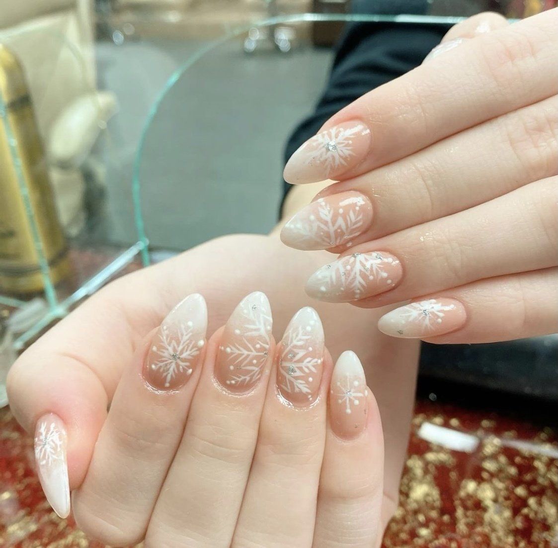 Hands with almond-shaped nails featuring a white snowflake design and ombre white to nude color.