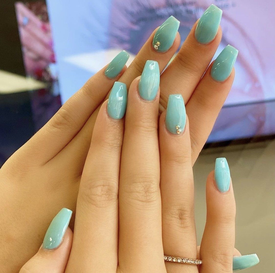 Hands with long, light blue, coffin-shaped nails; one hand with a small gem on one nail, a ring on the ring finger.