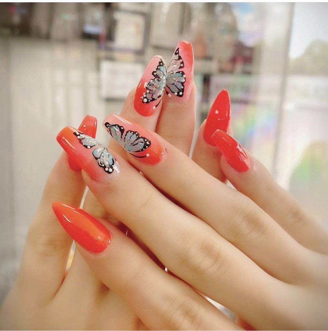 Woman's hand with long, red nails, two adorned with butterfly designs against a pink gradient.