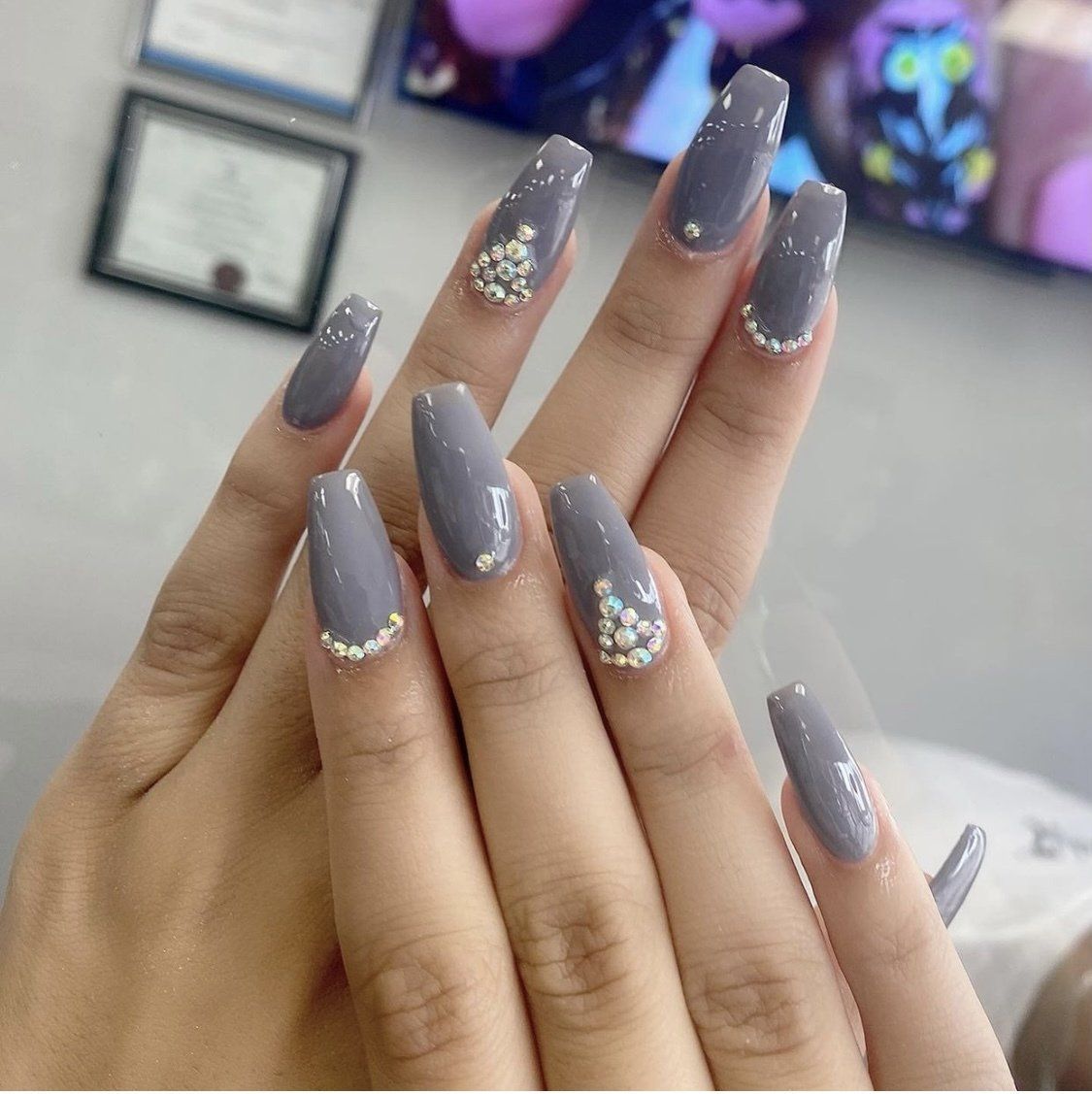 Hands with long, gray coffin-shaped nails, accented with small gems at the base of some nails.