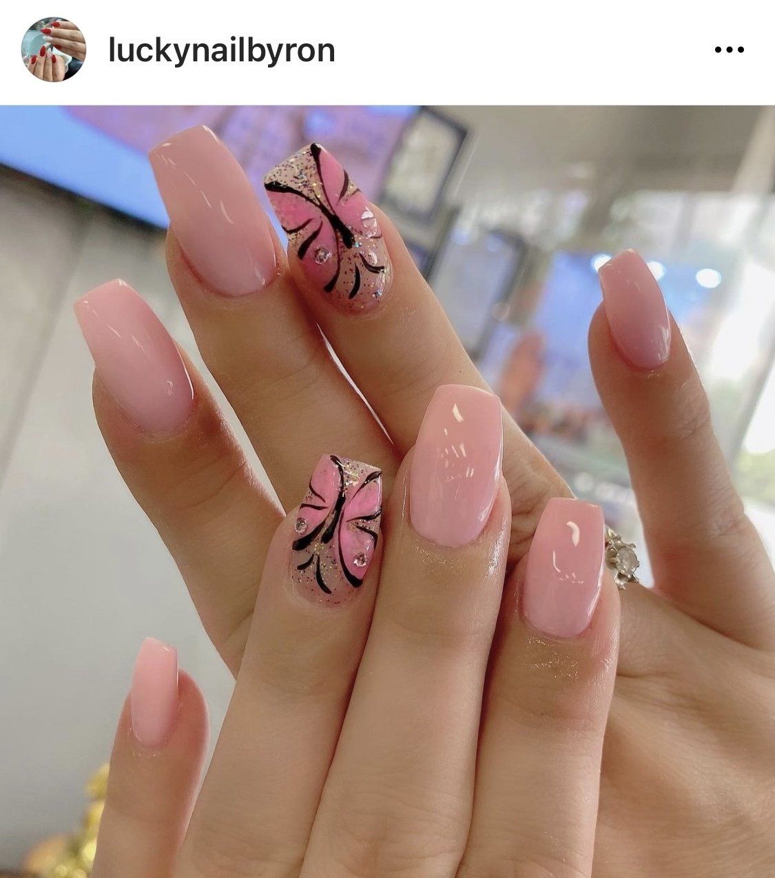 Pink coffin-shaped acrylic nails, two with black butterfly designs, on a hand in a salon.