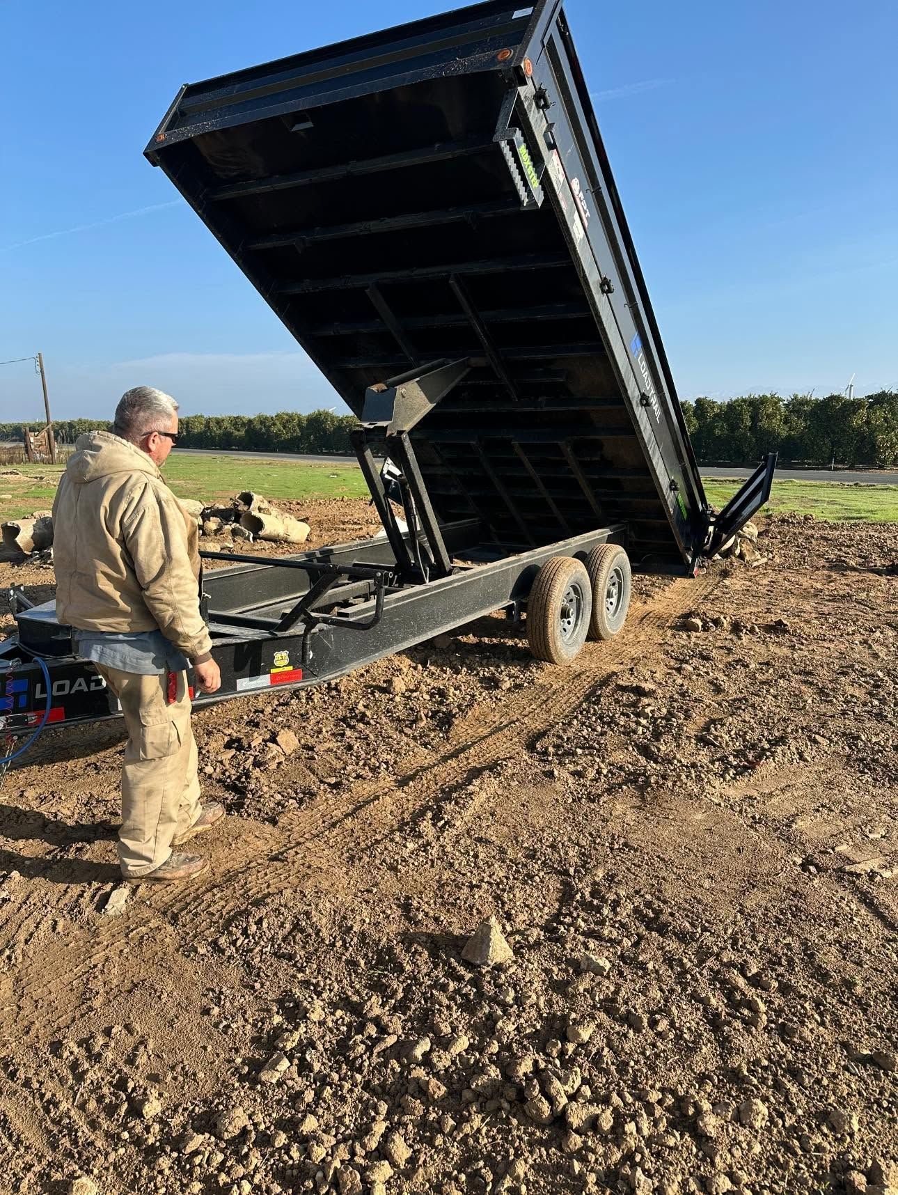 A man is standing next to a dump trailer in a dirt field.