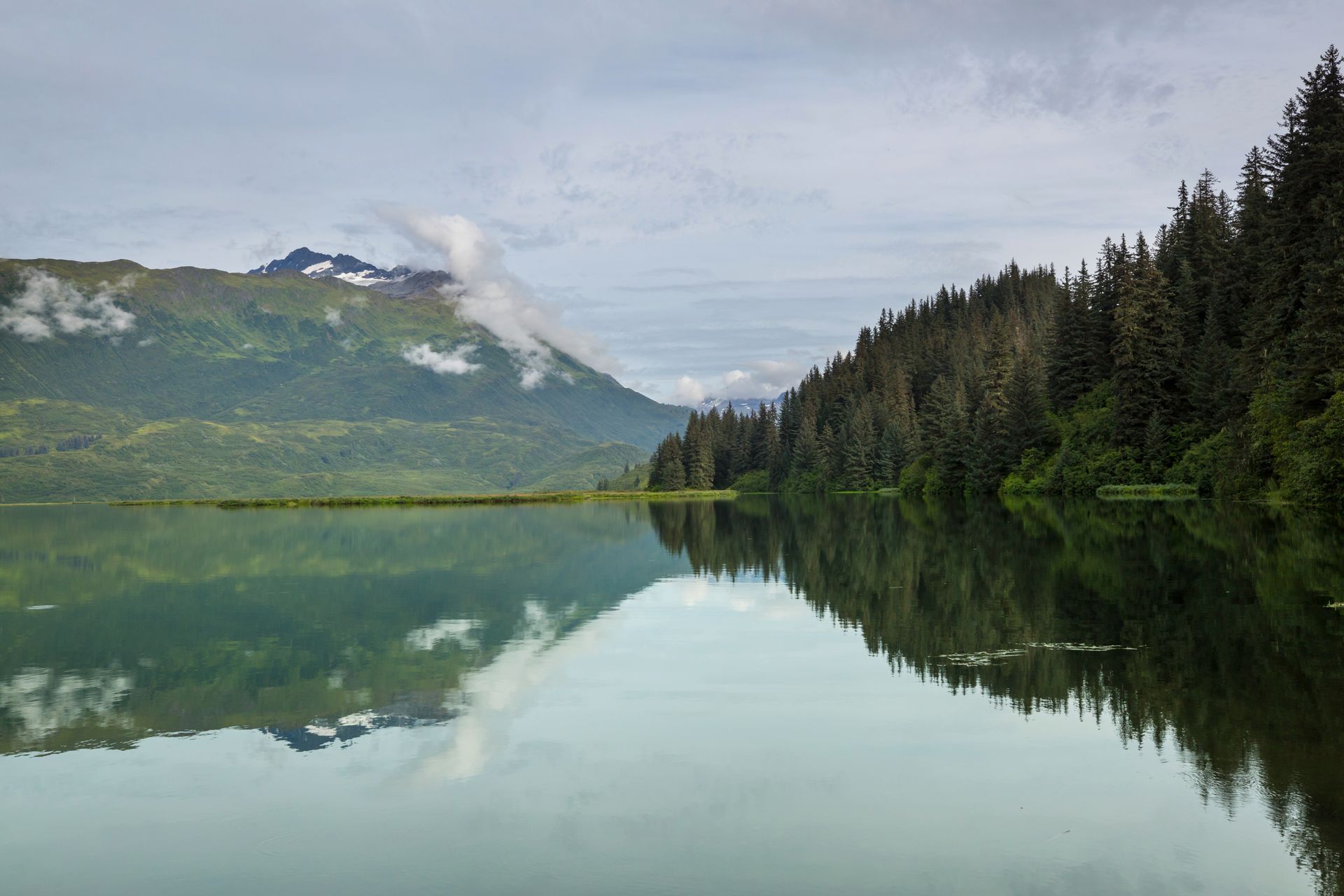 Calm lake reflecting forested hills and cloudy mountain.