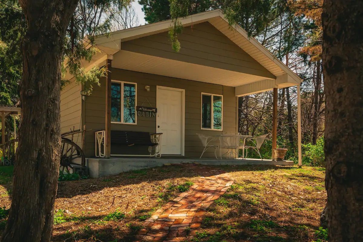 Small tan cabin with white trim, porch, and walkway surrounded by trees.