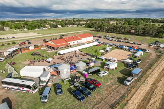 Aerial view of an outdoor event. People gathered near a large building, tents, and parked vehicles on a grassy area.