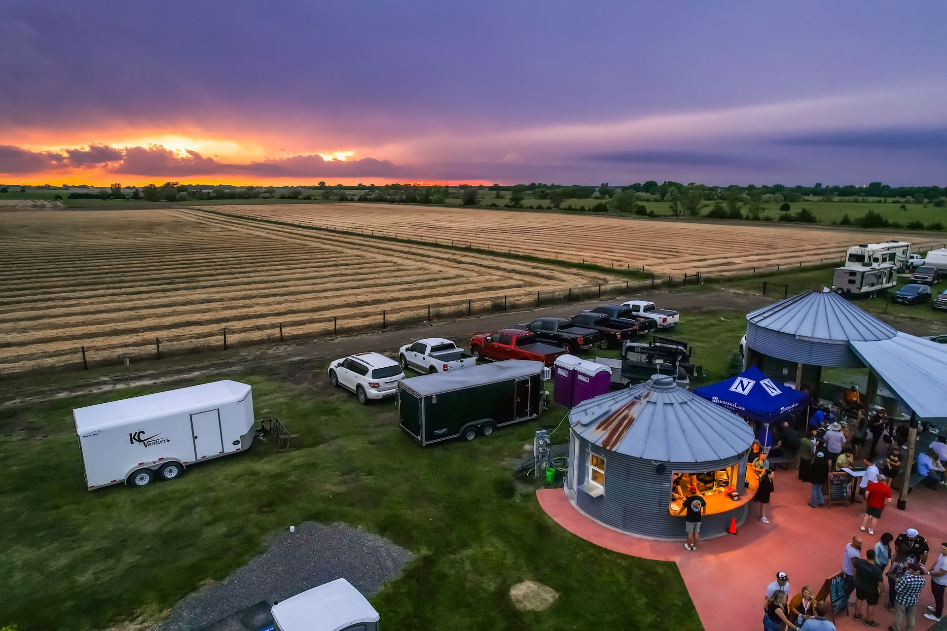 Aerial view of a farm event with silos, tents, cars, and a colorful sunset. People are gathered.