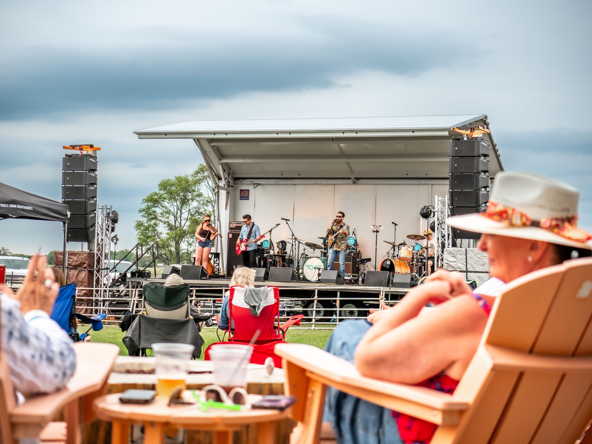 Band performing on outdoor stage; audience in chairs watches. Cloudy sky.
