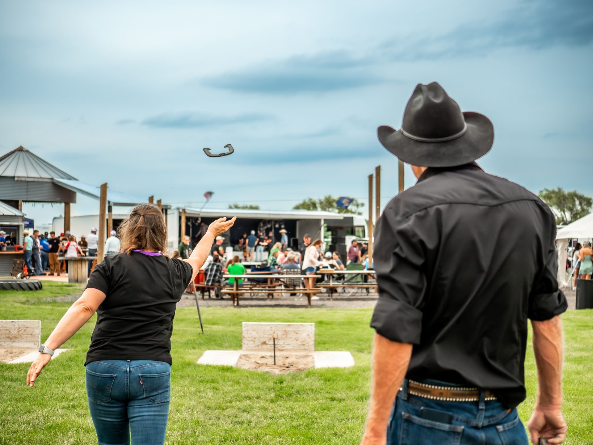 Woman throws a horseshoe, man in cowboy hat watches. Outdoor setting with onlookers.