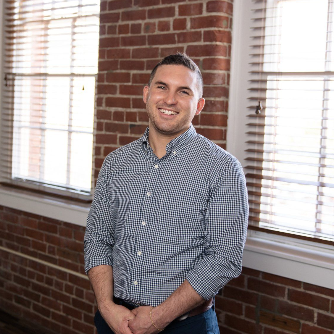 Smiling man in a checkered shirt standing in front of a brick wall and windows.
