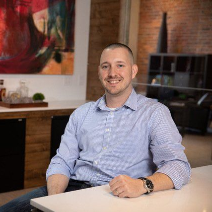 Man seated at a desk in a modern office, smiling with a laptop nearby.