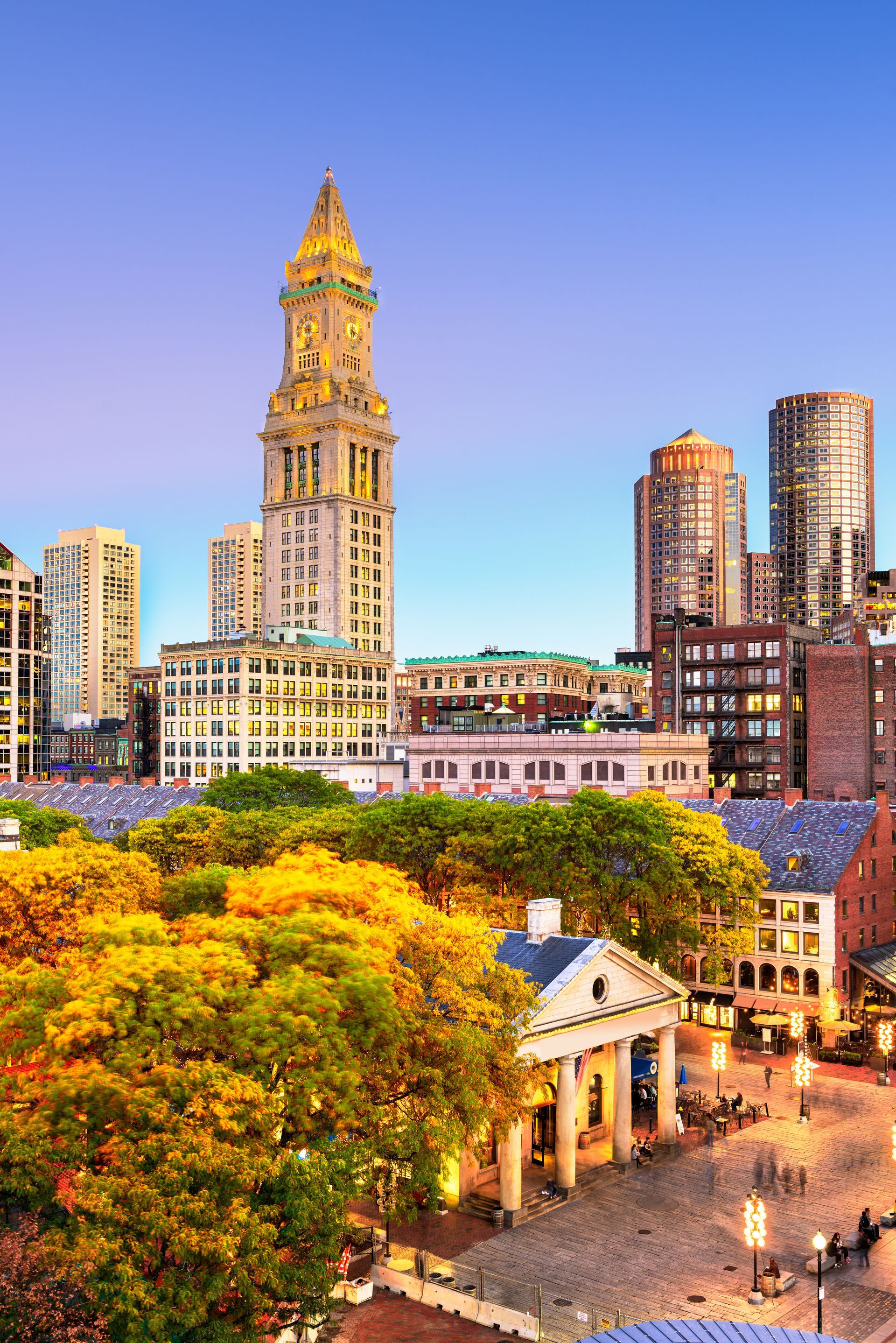 Sunset city skyline with a tall clock tower, colorful buildings, and a park plaza below