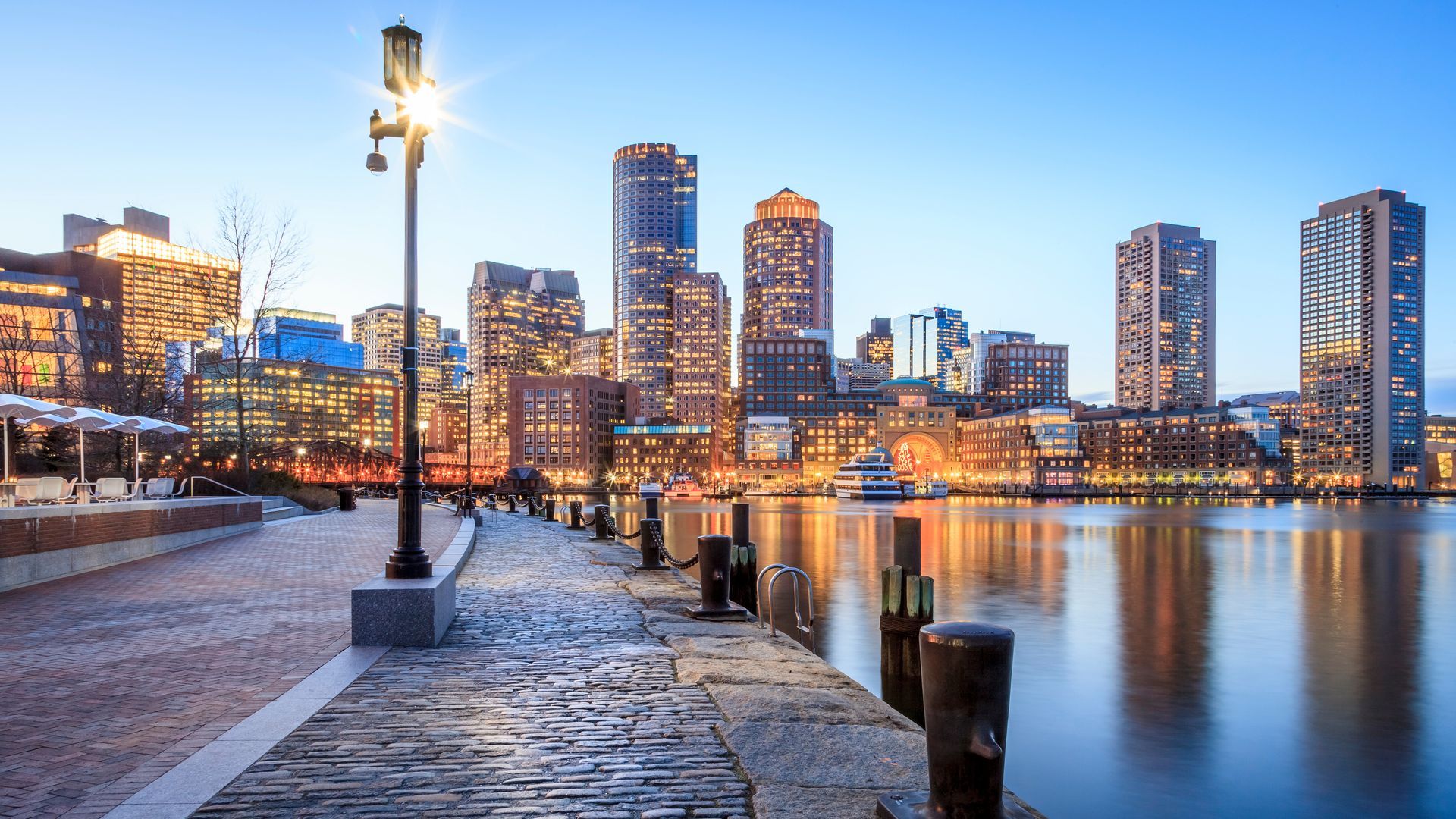 City skyline at dusk reflected on calm water beside a waterfront promenade and lamppost.