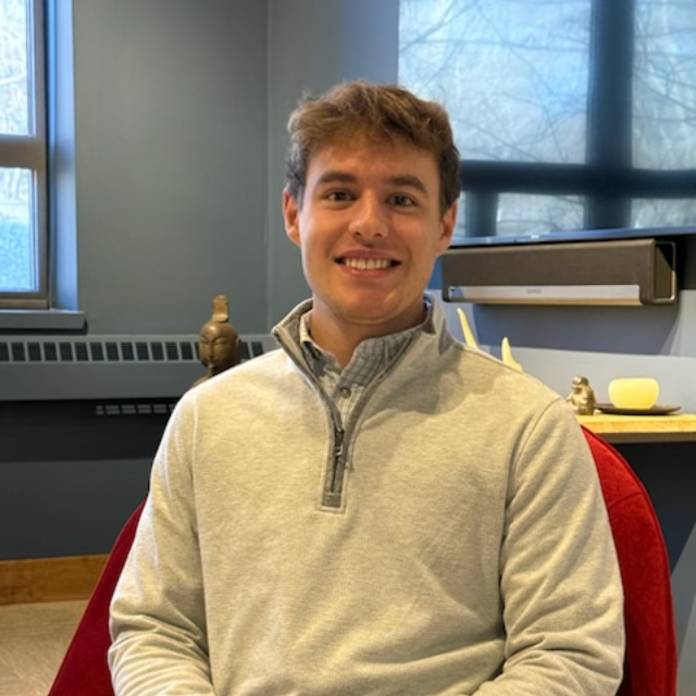 Smiling person in a light gray pullover seated indoors by a window and desk