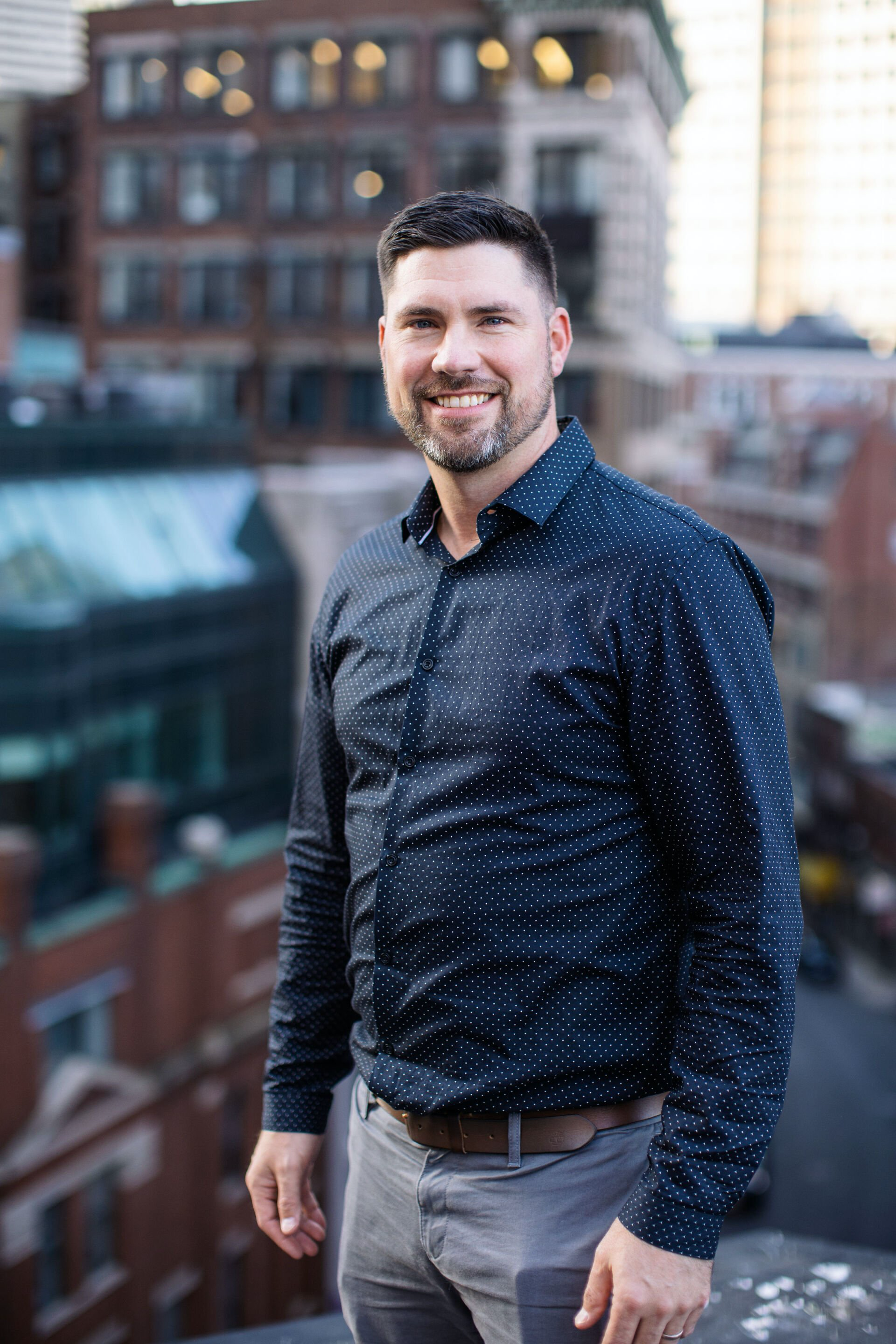 Smiling man in a navy patterned shirt standing on a rooftop with city buildings in the background