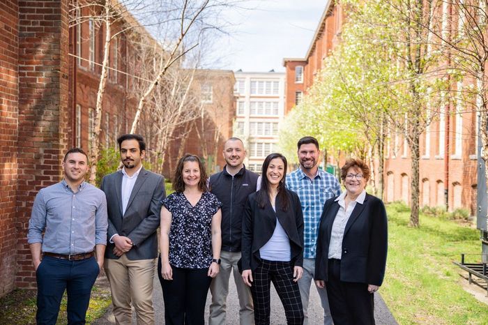 Six people standing outdoors on a sunny campus walkway between brick buildings.