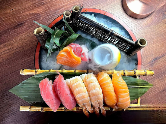 Piatto di sushi con nigiri, maki e guarnizioni colorate su foglie di banana e un tavolo di legno.