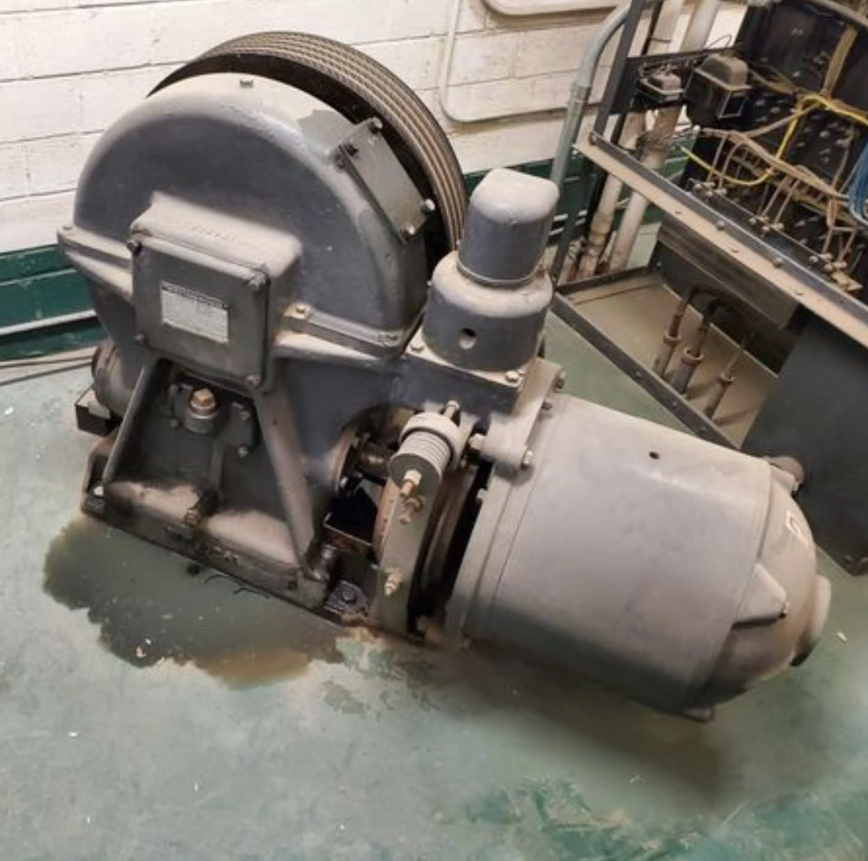 Grey machinery, possibly elevator equipment, with a motor and large wheel.