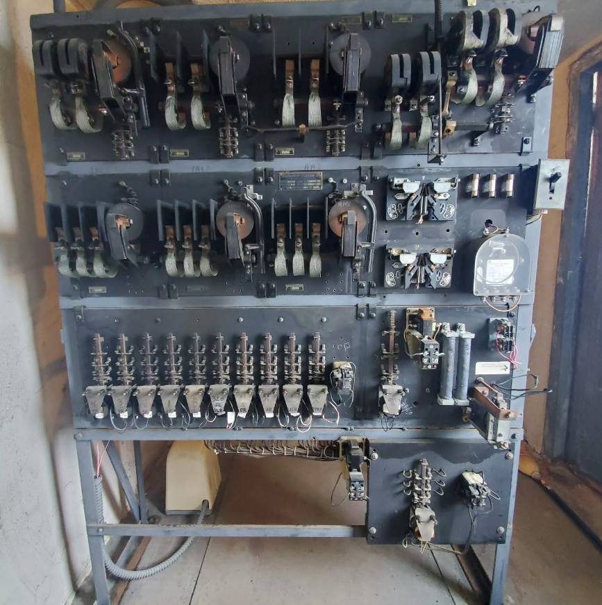An aged electrical control panel with various switches, dials, and components, mounted on a metal frame.