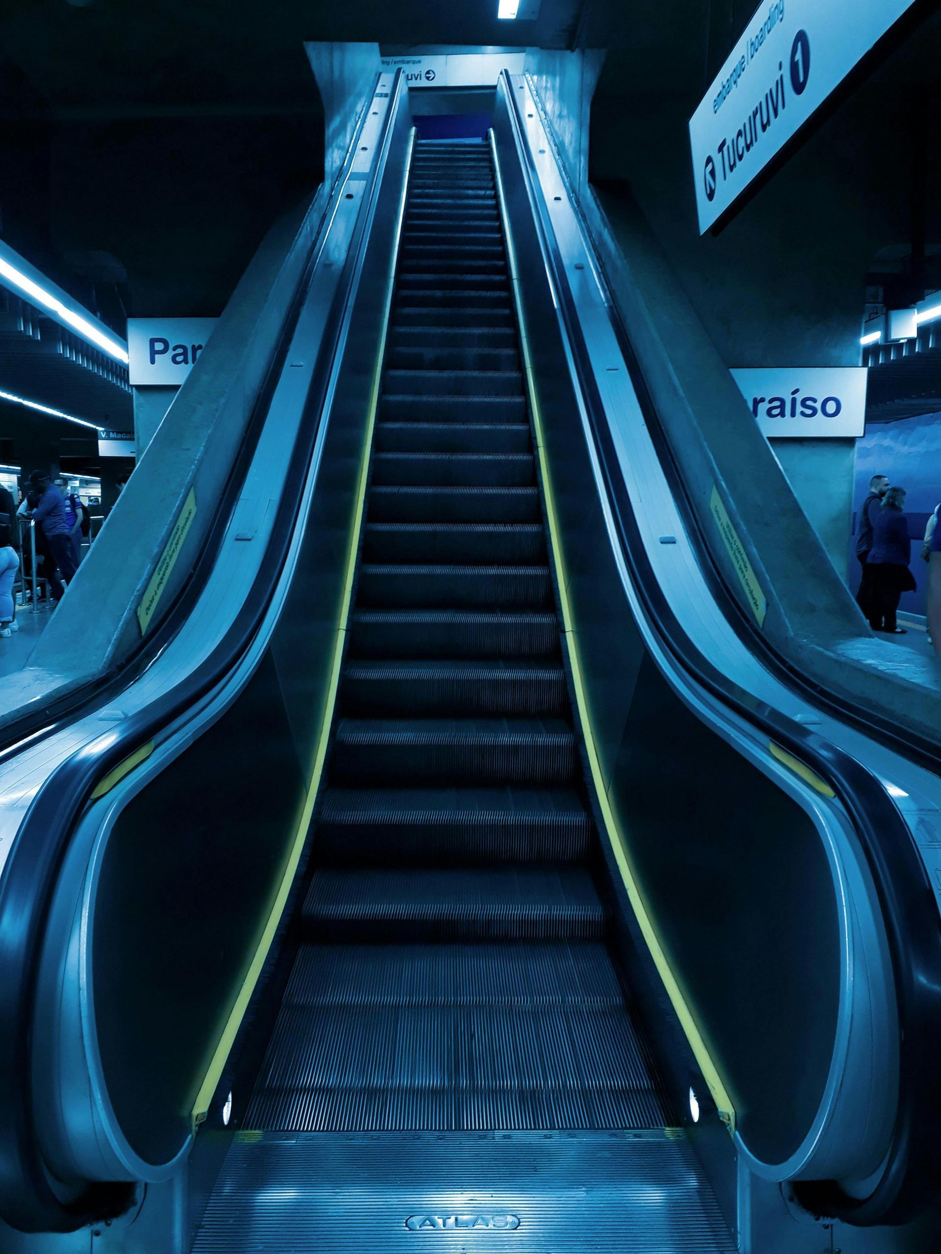 Escalator leading upwards in a blue-lit subway station. Yellow light lines the sides.