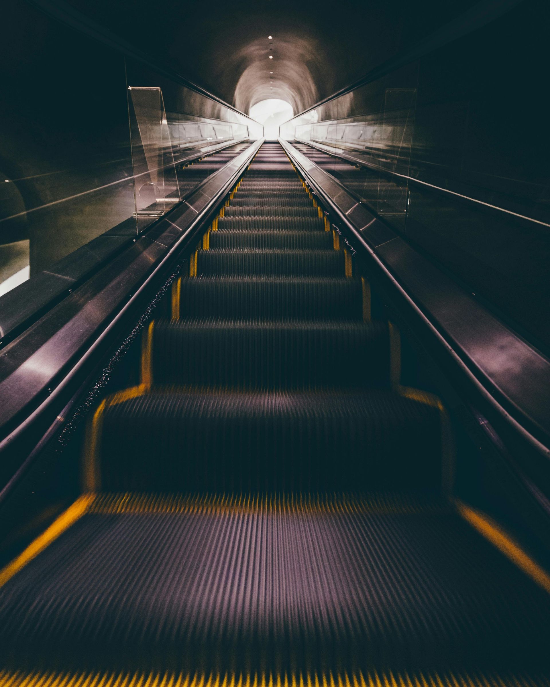Escalator ascending into a bright tunnel, with yellow safety lines, in a dark, metallic environment.